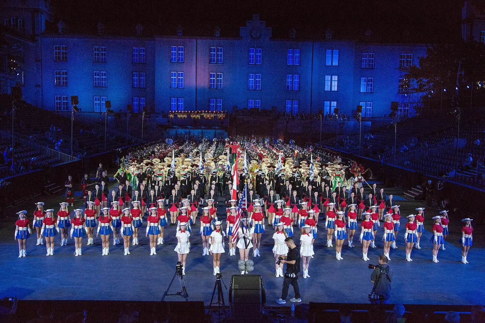 Marching bands and dancers fill the lit arena of the Basel Tattoo with the barracks facade behind