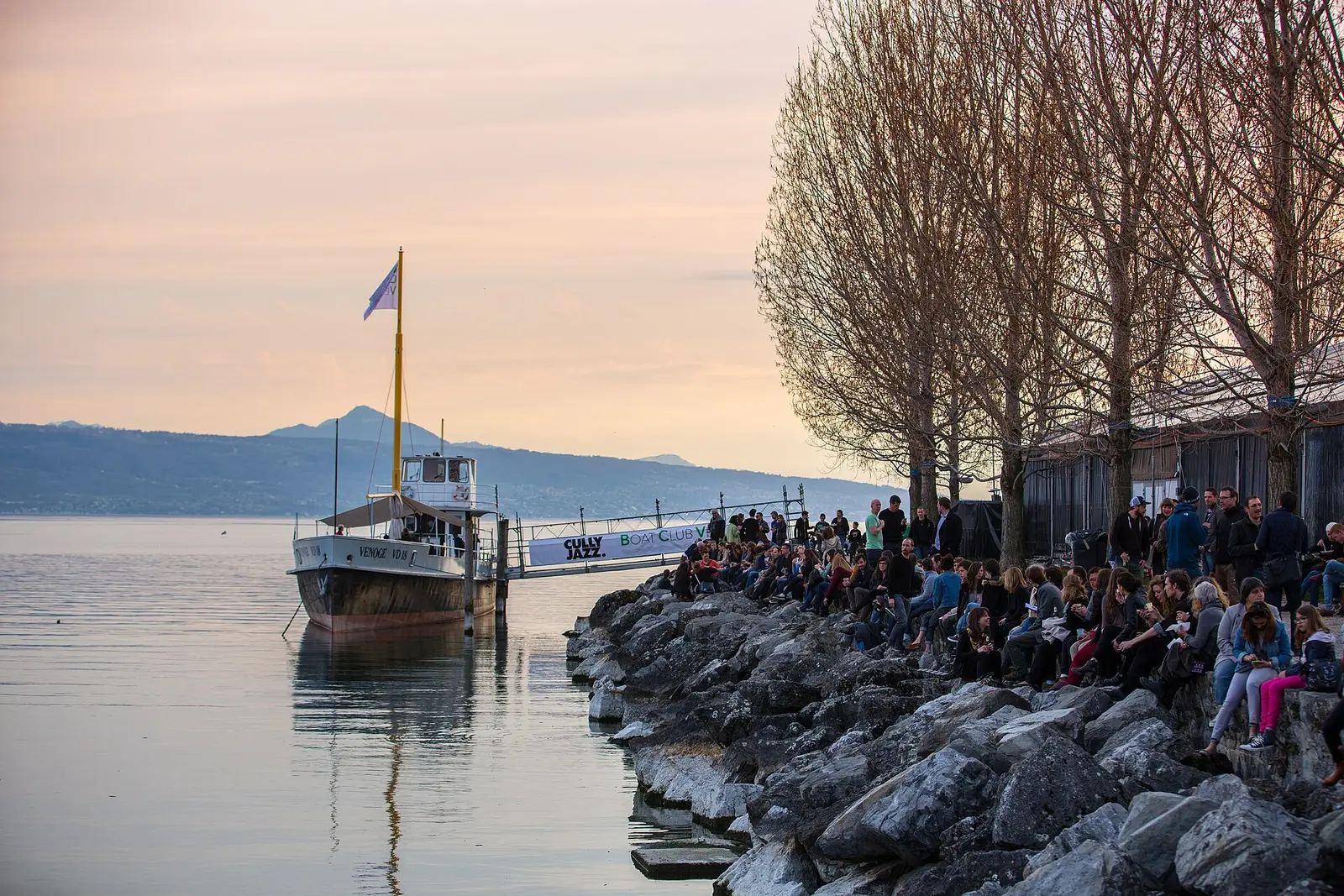 Audience seated on lakeside rocks at dusk beside a lake steamer at Cully